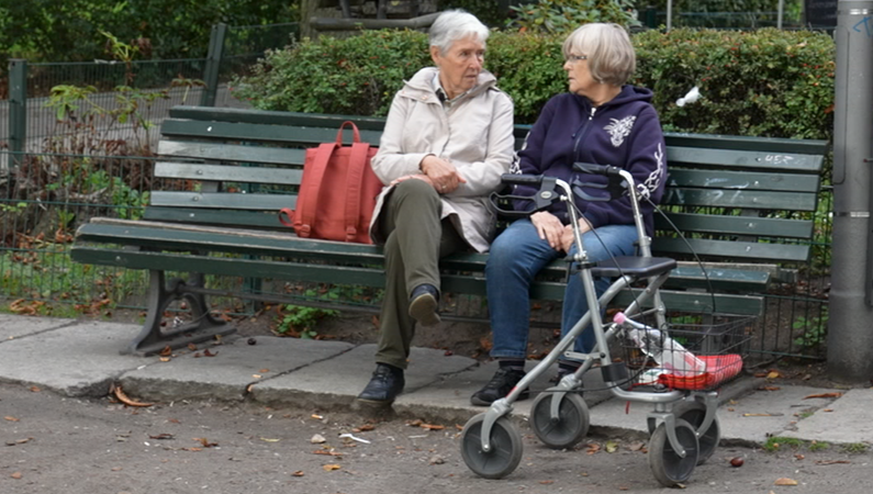 Zwei Seniorinnen auf einer Parkbank sitzend im Gespräch, mit Rollator