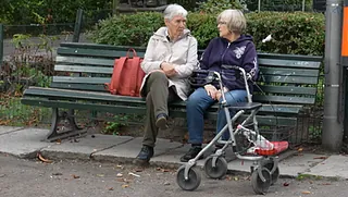 Zwei Seniorinnen auf einer Parkbank sitzend im Gespräch, mit Rollator